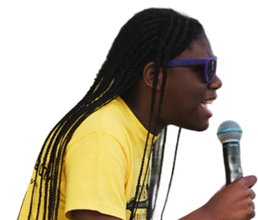 Person with long braided hair wearing purple sunglasses and a yellow shirt speaking or singing into a handheld microphone.